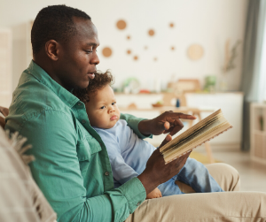 A dad sits with baby on his lap reading from an open book