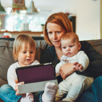 A woman with red hair holds an infant on her lap and has a toddler next to her on the couch and all are reading a tablet computer