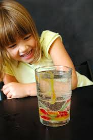 A girl smiles at a glass of liquid with a suspended gummy worm in it