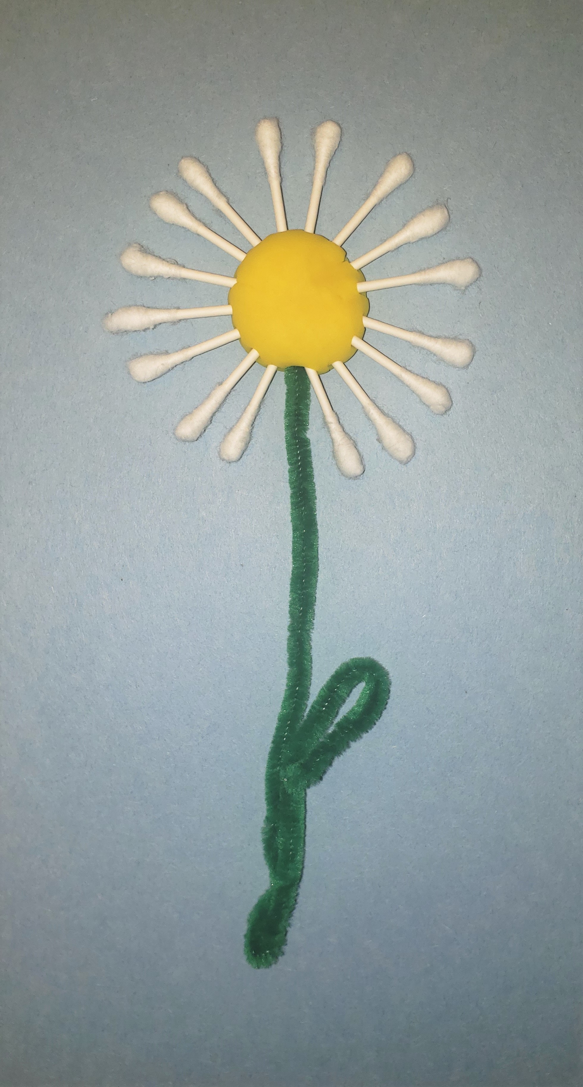 Daisy craft made from a yellow Play-doh circle surrounded by white cotton swabs, with a green pipe cleaner stem