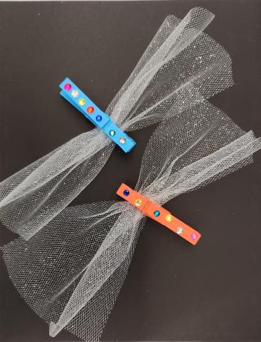 One orange and one blue clothespin, decorated with rhinestones, clasping pieces of sparkly tulle fabric to make dragonflies.
