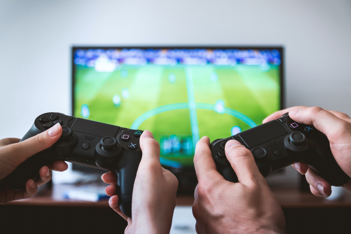 Two pairs of hands holding game controllers in front of a screen displaying a sports video game.